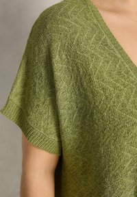 Camiseta de punto texturizado verde con mangas cortas y sueltas y escote en V en el torso superior y el brazo de una persona.