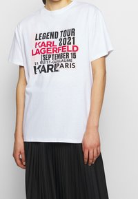 Bílý bavlněný tričko s grafickým potiskem a černým a červeným textem. Potisky zahrnují "Legend Tour", "Karl Lagerfeld" a detaily o Paříži.