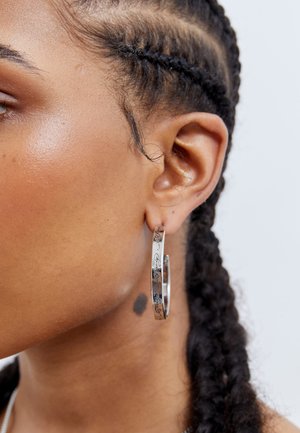 Srebrna obročasta uhana z vgraviranimi detajli, ki prikazujejo gladko površino in krožno obliko, nosijo se na ušesu ter izpostavljajo zapleteno zasnovo.