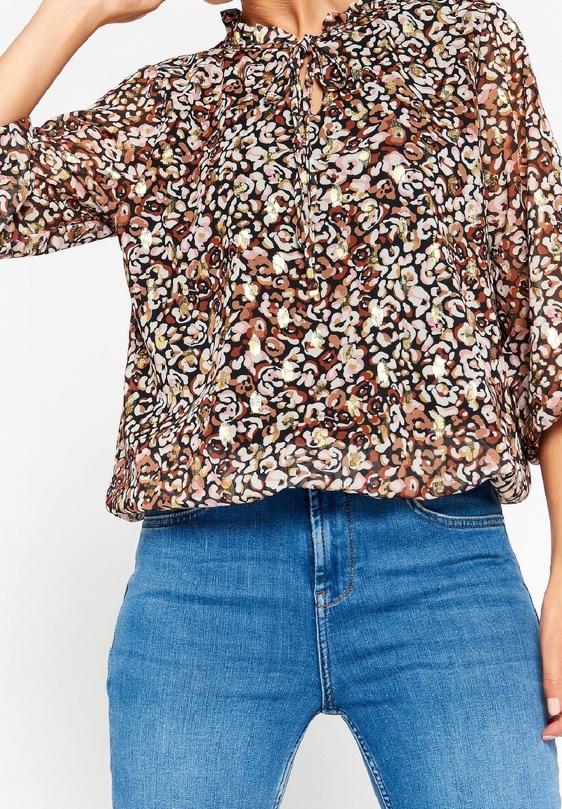 Femme portant une blouse en voile à manches longues avec un imprimé léopard multicolore et des accents dorés, associée à un jean bleu taille haute.