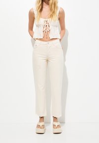 Bílý pletený crop top s volnými rukávy a předním zavazováním; zkombinovaný s světle béžovými džíny s vysokým pasem, které mají třepené lemy a jemné opotřebení.