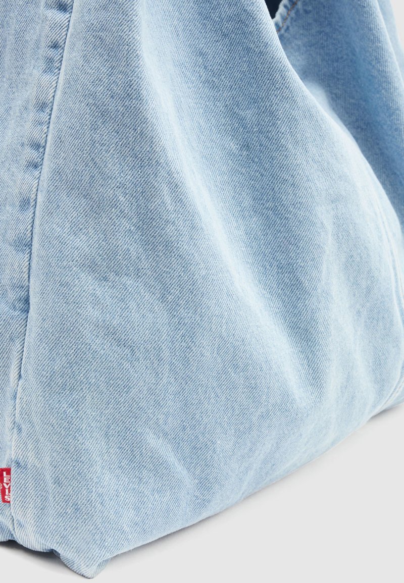 Angolo di una borsa tote in denim azzurro con cucitura visibile e etichetta rossa con il logo Levi's su sfondo bianco.