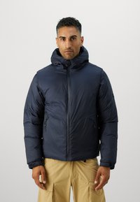 Veste matelassée bleu marine avec un col haut et une capuche. Dotée d'une fermeture éclair à l'avant et de poches latérales zippées, d'une texture lisse et d'une silhouette ajustée.