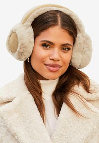 Auriculares de piel sintética beige con una textura suave y un diseño curvado; cuentan con una banda suave a juego para mayor comodidad. Los lleva puesta un modelo con un abrigo cálido.