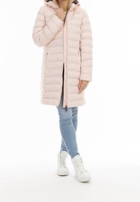 Žena v světlém růžovém dlouhém péřovém kabátu, světlých modrých džínách a bílých teniskách s vysokým svrškem, stojí s překříženýma nohama a rukou u límce.