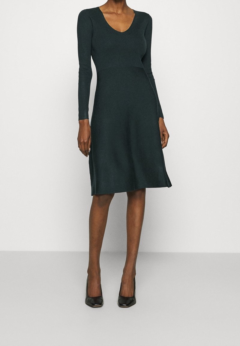 Femme portant une robe vert foncé à manches longues et longueur genou avec un décolleté arrondi, ainsi que des chaussures à talons noirs, sur un fond uni.