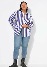 Camisa de botones a rayas moradas y blancas con dos bolsillos en el pecho, combinada con jeans ajustados azul claro y botas negras hasta el tobillo.