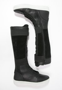 Černé kožené a semišové vysoké boty do kolen s texturovanými popruhy, zipovým uzávěrem a bílou gumovou podešví. Šněrovací design na dolní části.