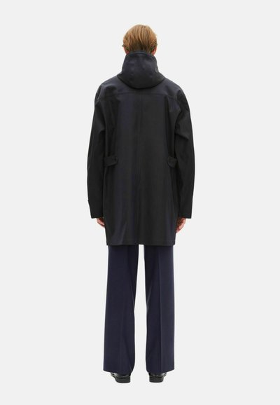 Manteau noir à capuche avec une coupe ample, doté de poches latérales et d'une texture lisse et résistant à l'eau. Associé à un pantalon large bleu marine.