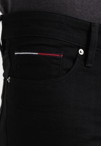 Svarta denimjeans har en tät passform med en silverfärgad knapp och en framficka som är accentuerad med röd, vit och blå sömnad.