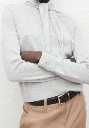 Personne portant un sweat à capuche gris clair et un pantalon beige avec une ceinture noire, les bras croisés, debout contre un mur blanc texturé.