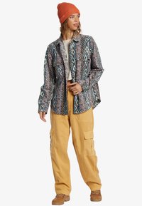 Patterned shirt in blue, cream, and rust with abstract designs; worn over a white top, paired with yellow cargo pants and brown boots.