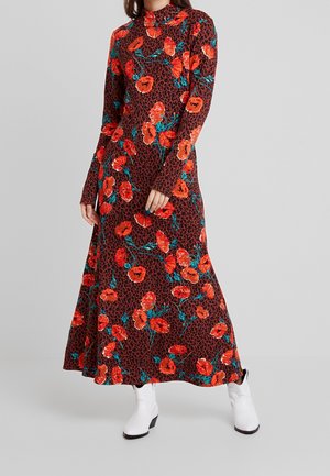 Kvinde iført en langærmet, gulvlængde brun kjole med rødt blomstermønster og sort leopardprint, kombineret med hvide ankelstøvler.