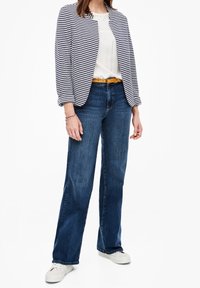 Blazer rayé bleu marine et blanc porté sur un haut blanc, associé à un jean bleu et des baskets blanches. Une ceinture jaune apporte une touche contrastante.