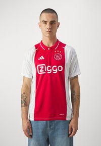 Maillot de football Ajax avec un devant rouge et des manches, un col et des accents blancs. Affiche le logo "Ziggo" et l'emblème du club, fabriqué en tissu respirant.