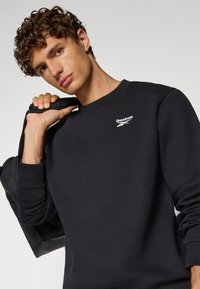 Czarna bluza z okrągłym dekoltem i ściągaczami, z białym logo Reebok na piersi, trzymana na ramieniu z czarną torbą.