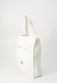 Vit canvas shoppingväska med en zip-försedd framficka, prydd med en liten broderad design på sidan. Robust tyg och platt botten.