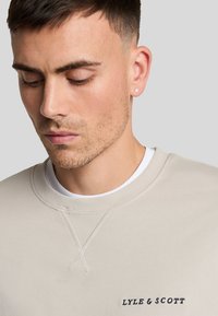 Sudadera beige con cuello redondo, que presenta un logotipo negro en contraste de "LYLE & SCOTT" en el lado izquierdo del pecho y detalles de costura sutiles.