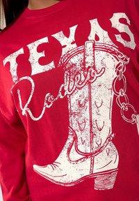 Sudadera de algodón roja con un gráfico blanco de una bota de vaquero con una cadena, el texto dice "Texas Rodeo". La textura desgastada añade detalle.