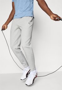 Lys grå sweatpants lavet af et blødt materiale, der har en smal pasform og et lille logo. Bæreren hopper med sjippetov iført hvide sneakers.
