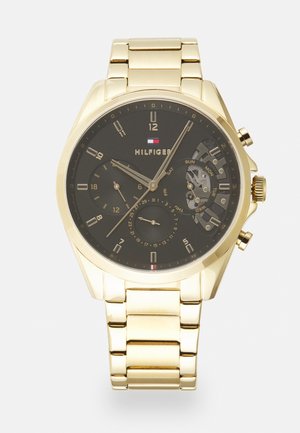 Montre-bracelet en métal doré avec cadran noir, trois sous-cadrans pour la date, le jour et l’heure 24h, et logo Tommy Hilfiger au centre.