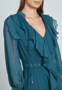 Teal Kleid mit gerafftem Ausschnitt und langen durchsichtigen Ärmeln. Geraffte Taille mit einem Band und goldfarbenen Akzenten. Leichtes, strukturiertes Material.
