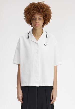 Jeune personne aux cheveux bouclés portant une chemise blanche à manches courtes avec un bord noir au niveau du col et un pantalon noir, sur un fond uni.