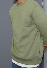 Felpa verde oliva con colletto a girocollo, polsini e orlo a coste. Presenta un piccolo logo sul petto, indossata sopra una camicia bianca.