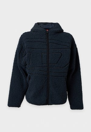 Jeune homme portant une veste polaire noire sur un t-shirt blanc graphique, un pantalon large noir et des baskets noires et argentées, debout sur un fond uni.
