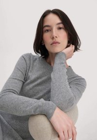 Maglione grigio lavorato a maglia con colletto rotondo e polsini a coste. Il modello posa con la mano sul mento, seduto su una superficie morbida di colore chiaro.