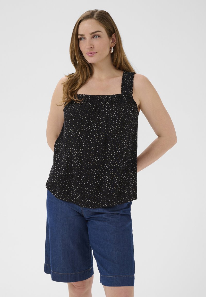 Jeune femme aux longs cheveux bruns portant un haut noir sans manches à petits pois blancs et un short en denim bleu jusqu'aux genoux.