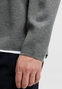 Desna ruka opuštena sa strane, nosi sivi džemper s dugim rukavima preko bijele košulje i tamne hlače na jednostavnoj podlozi.