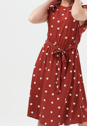 Frau in einem ärmellosen roten Kleid mit weißen Polka-Dots und einem gebundenen Taillengürtel, die vor einem schlichten Hintergrund ihre Haare richtet.
