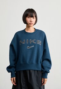 Sötétkék, túlméretezett pulóver kerek nyakkal és bordás mandzsettákkal. Jellemzi a nagy "NIKE" logó, kontrasztos fehér és szürke szöveggel.