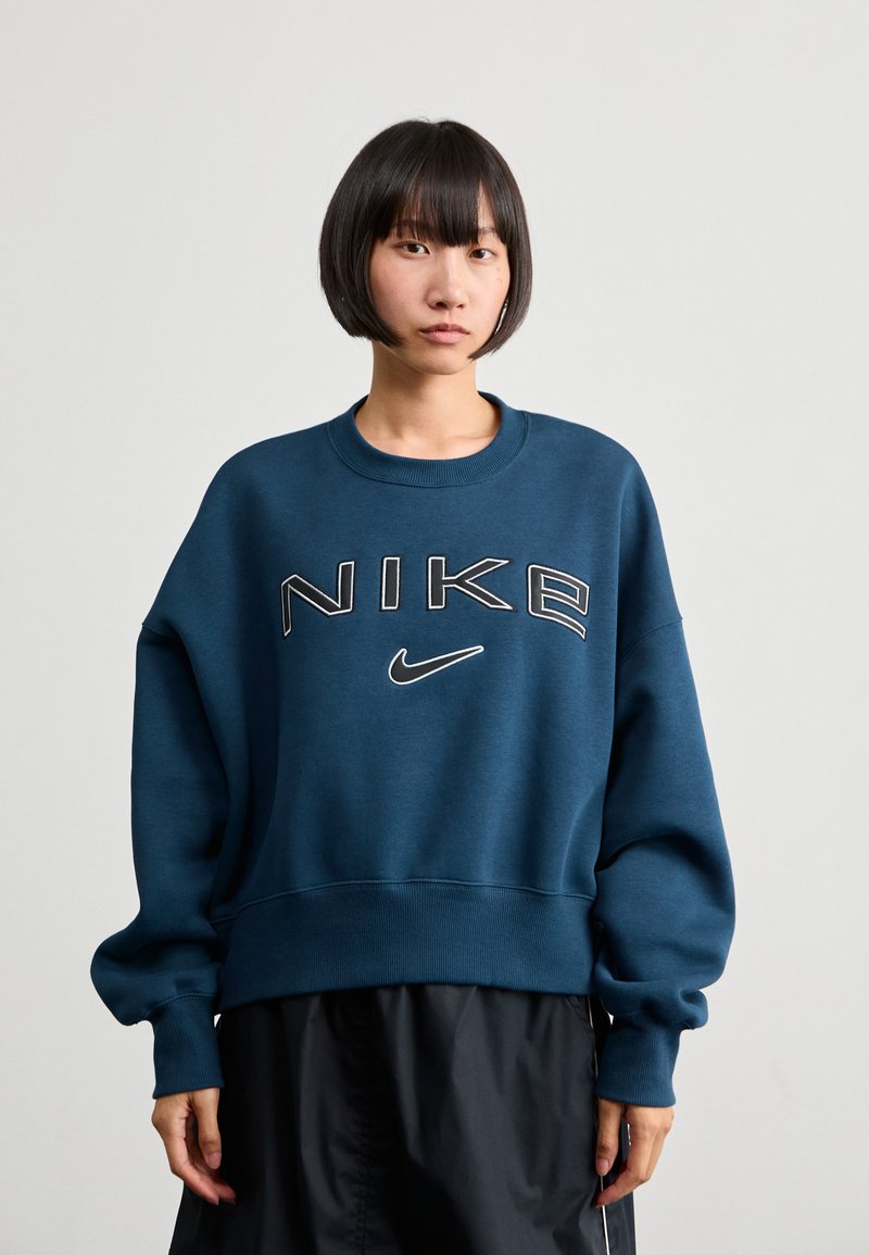 Sötétkék, túlméretezett pulóver kerek nyakkal és bordás mandzsettákkal. Jellemzi a nagy "NIKE" logó, kontrasztos fehér és szürke szöveggel.