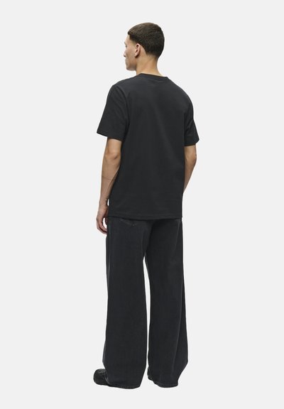 Tricou negru din bumbac cu guler rotund și mâneci scurte, purtat cu blugi negri cu croi larg. Design simplu, fără modele sau logo-uri vizibile.