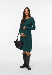 Knälång, långärmad grön stickad klänning; figurnära liv, lös kjol. En svart läderhandväska och knähöga svarta stövlar fulländar looken.