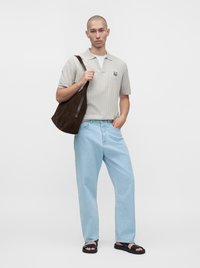 Hombre con polo de manga corta gris claro, vaqueros azul claro, sandalias negras y llevando un gran bolso de hombro marrón oscuro.