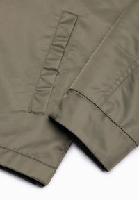 Gros plan sur la manche d'une veste vert olive et une poche à ouverture passepoilée, montrant la texture du tissu et les détails de couture.