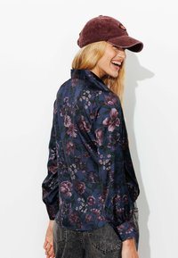 Blouse florale en bleu foncé et violet, avec de longues manches bouffantes et un poignet à boutons. Assortie à une casquette en velours côtelé bordeaux.