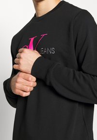 Sweatshirt noir avec un col rond et des manches longues. Présente des logos brodés roses et gris proéminents sur la poitrine. Texture en coton.