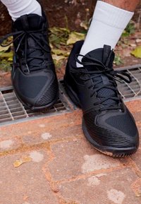 Schwarze sportliche Sneaker mit strukturiertem, synthetischem Obermaterial, ausgestattet mit Mesh-Einsätzen, Schnürdesign und Gummisohle, auf einer befestigten Oberfläche gezeigt.