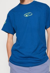 Modré bavlněné tričko s kulatým výstřihem, na hrudi s žlutozeleným logem Nike a kontrastním akcentem na rukávu.