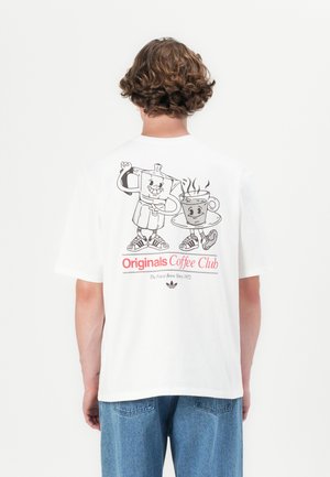 Hvid T-shirt med grafisk tryk af en smilende kaffekande og kop. Teksten siger "Originals Coffee Club" i rødt, med blå jeans synlige.