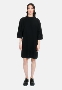 Črna oversized obleka z visokim ovratnikom in rokavi do tri četrtine, narejena iz gladkega materiala, v kombinaciji z svetlečimi črnimi čevlji.