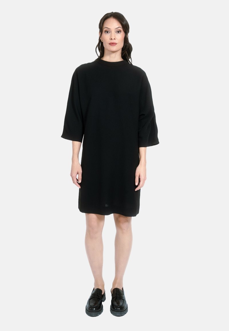 Črna oversized obleka z visokim ovratnikom in rokavi do tri četrtine, narejena iz gladkega materiala, v kombinaciji z svetlečimi črnimi čevlji.