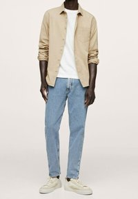 Chemise beige boutonnée, t-shirt blanc, jean bleu clair, et baskets crème. Tenue décontractée avec une coupe structurée et une texture en coton.