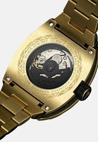 Goudkleurig horloge met een roestvrijstalen band. Beschikt over een zichtbare mechanische beweging en verfijnde gravures op de achterzijde van de behuizing.