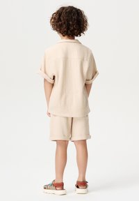 Beige, textuurde, korte mouwen shirt met opgerolde mouwen, gecombineerd met bijpassende shorts. Beide kledingstukken hebben een relaxte pasvorm. Casual sandalen maken de look compleet.