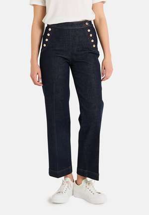 Pantalones de mezclilla oscura de cintura alta con pierna recta, que presentan grandes botones dorados en el costado y costuras en contraste, combinados con zapatillas blancas.